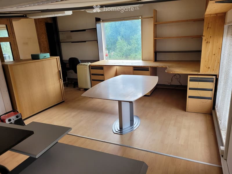 A modern, well-lit office space with wooden flooring, large windows, a central round table, chairs, cabinets, and shelves.