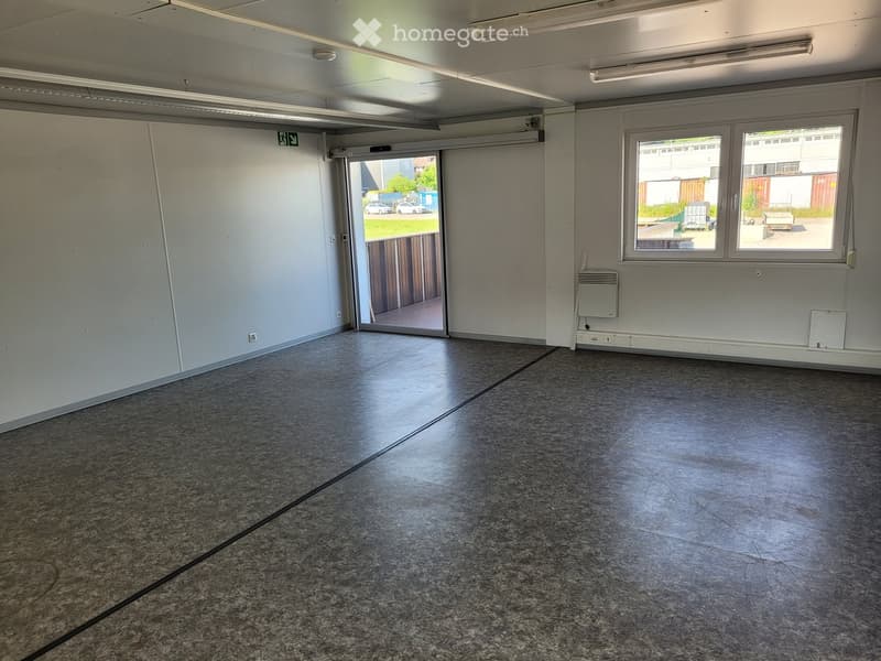 Empty room with grey walls, grey tiled floor, large windows, and a sliding door leading to an outdoor area.