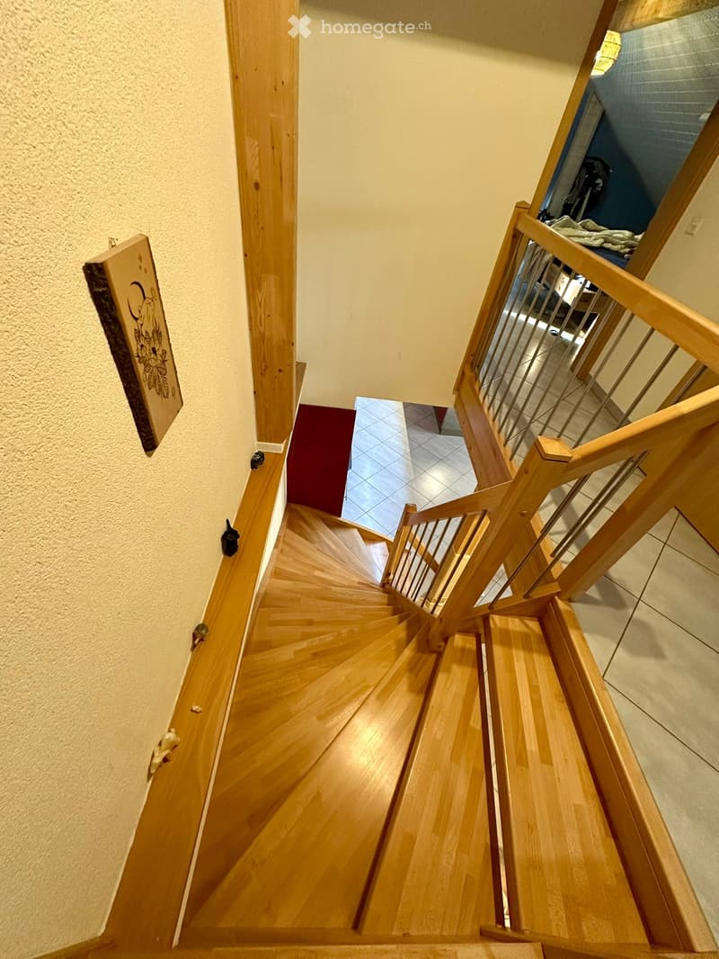 Spiral wooden staircase with metal railing, beige walls, and a red carpeted landing leading to an adjacent room.
