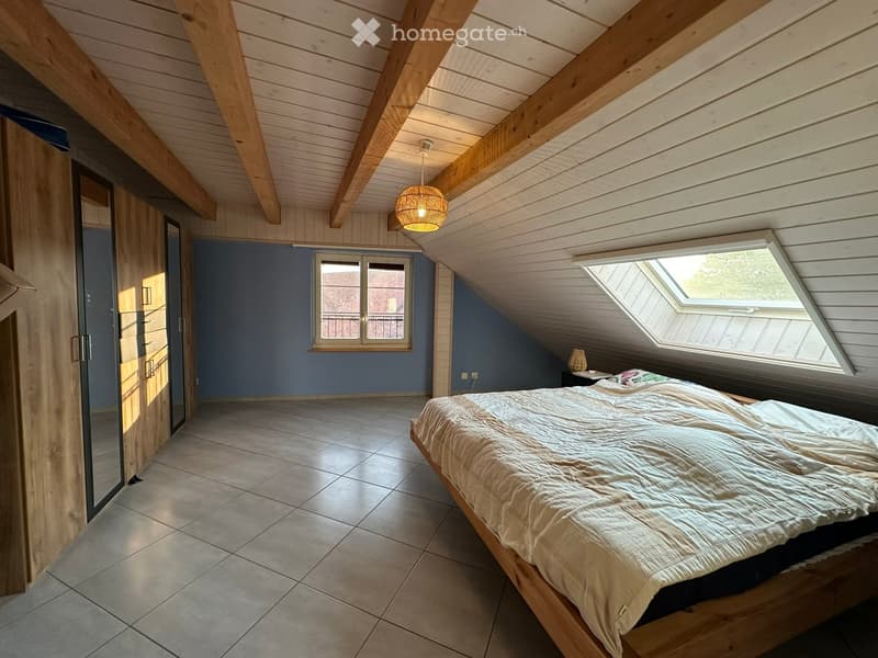 Bright attic bedroom with wooden beams, tiled floor, large windows, and a bed with white bedding.