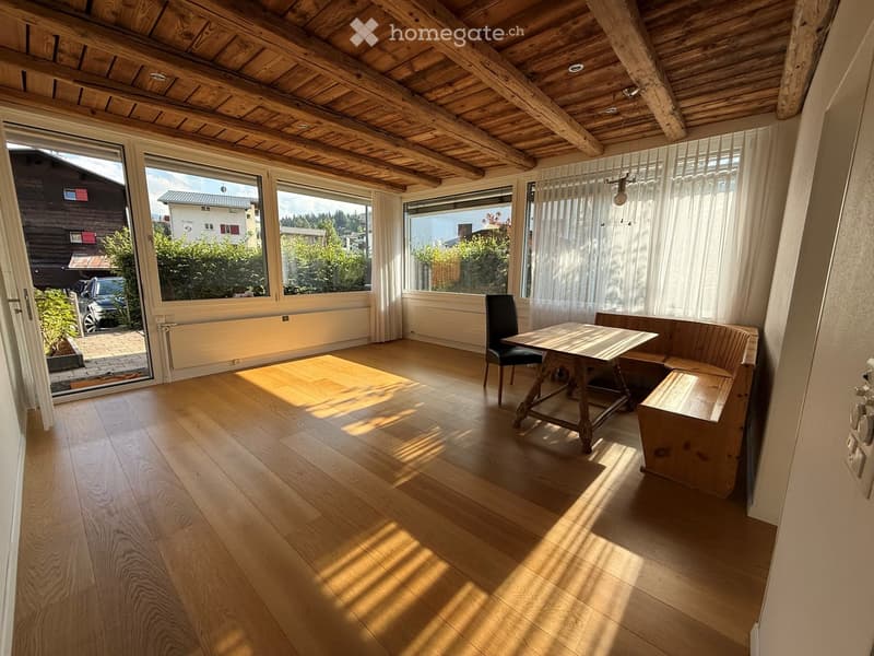 Bright room with wooden floor, large windows, a wooden table, a chair, and a bench. The room has a wooden ceiling with exposed beams and a view of the outdoors.
