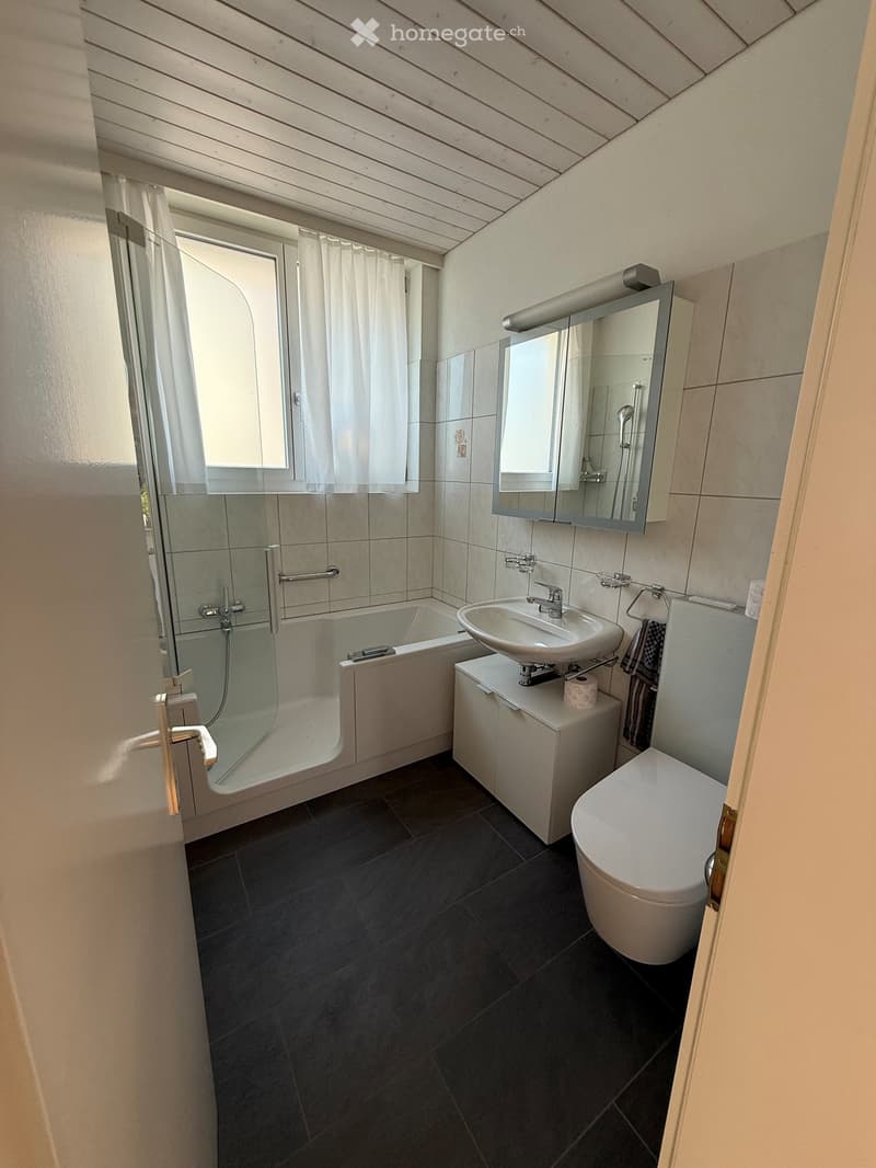 Modern bathroom with dark tiled floor, white bathtub, sink, toilet, mirrored cabinet, and a window with white curtains.
