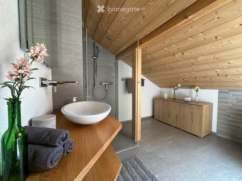 Modern bathroom with wooden ceiling, grey tiles, a large sink, a shower area, and decorative vases with flowers.