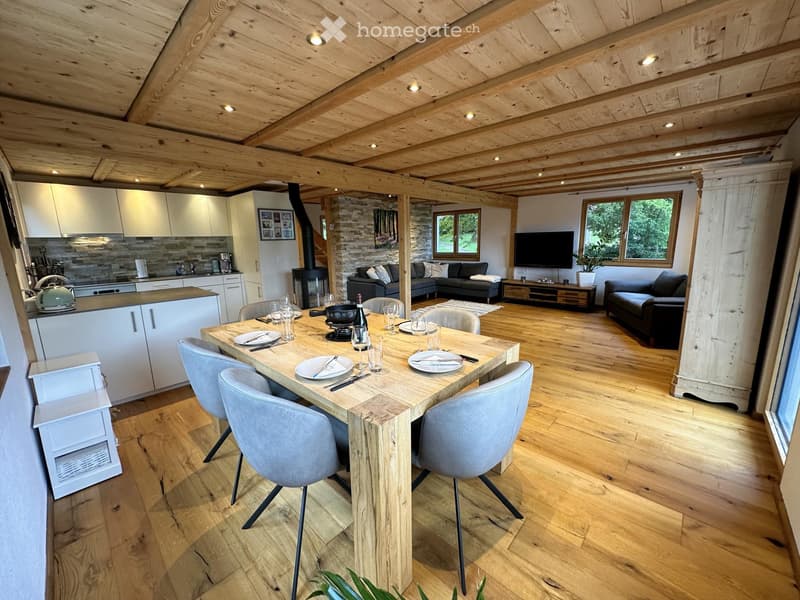 Open-plan living and dining area with exposed wooden beams, a dining table with chairs, a sofa, a television, and a modern kitchen with white cabinets and stainless steel appliances.