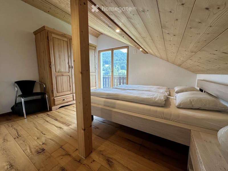 Cozy attic bedroom with wooden floors, slanted wooden ceiling, large window offering a scenic mountain view, a double bed, a wardrobe, and a black chair.