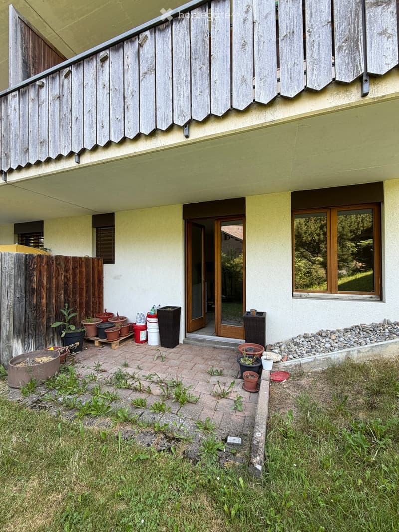 Outdoor patio area with brick tiles, potted plants, and a wooden fence. The entrance to the house is visible with a glass door and wooden shutters on the upper level.