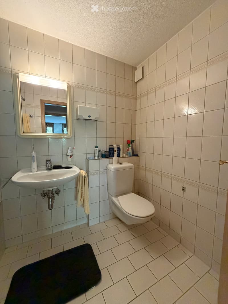 A small bathroom with a white tiled floor and walls, featuring a sink with a mirror, a toilet, a towel, and a bath mat.