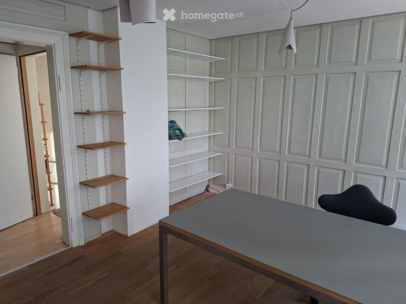A modern, minimalist room with wooden flooring, white paneled walls, built-in shelves, a grey table, and a black chair.