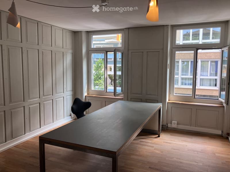 Minimalist room with wooden flooring, grey walls, large windows, a long dark table, and a single chair.