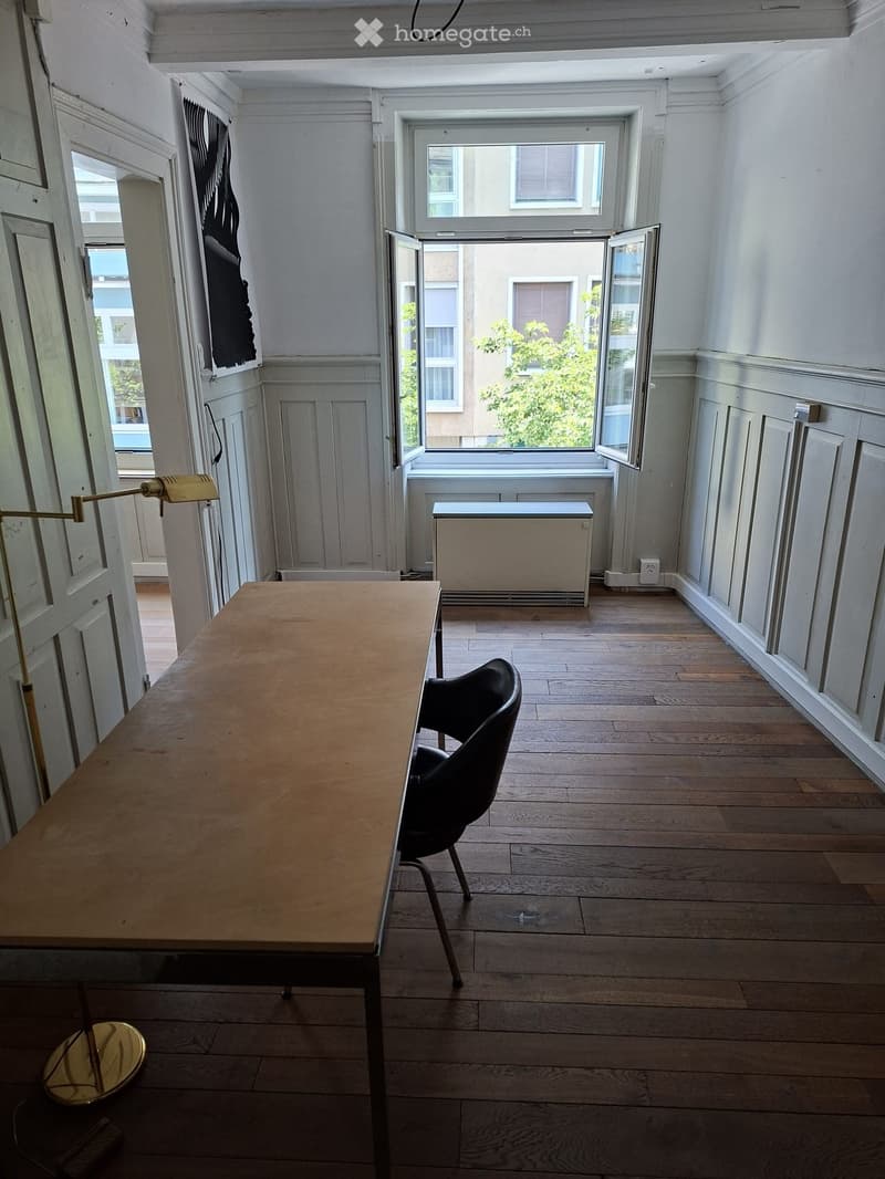 Bright room with wooden flooring, large windows, a table, a chair, and a wall-mounted lamp.