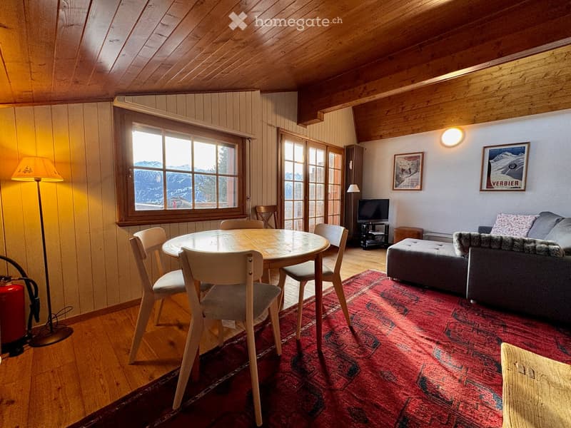 Cozy living room with wooden beams, exposed wooden floor, large window with mountain view, sofa, ottoman, dining table with chairs, television, and artwork on walls.