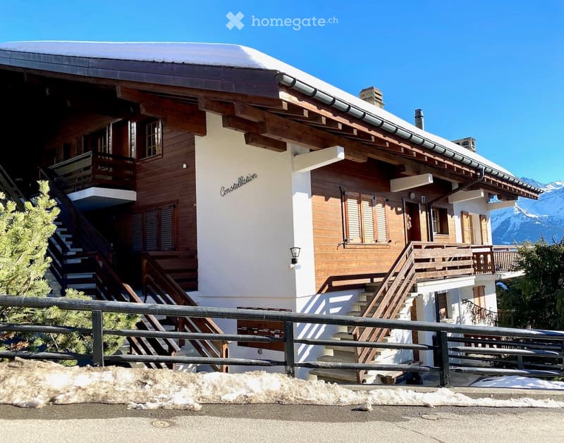 A large, multi-story wooden house with a snow-covered roof and balconies, situated in a mountainous area with snow on the ground.