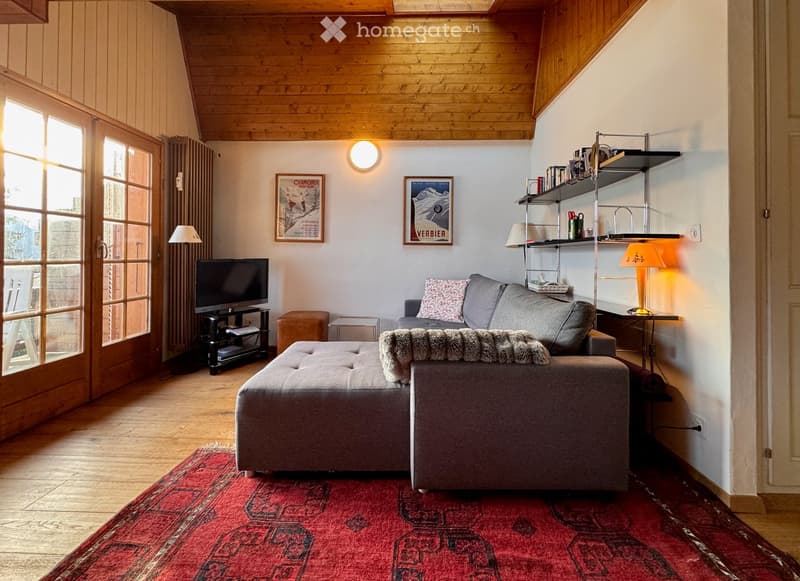 Cozy living room with wooden ceiling, large windows, grey sofa, ottoman, coffee table, television, floor lamp, shelves, and artwork.