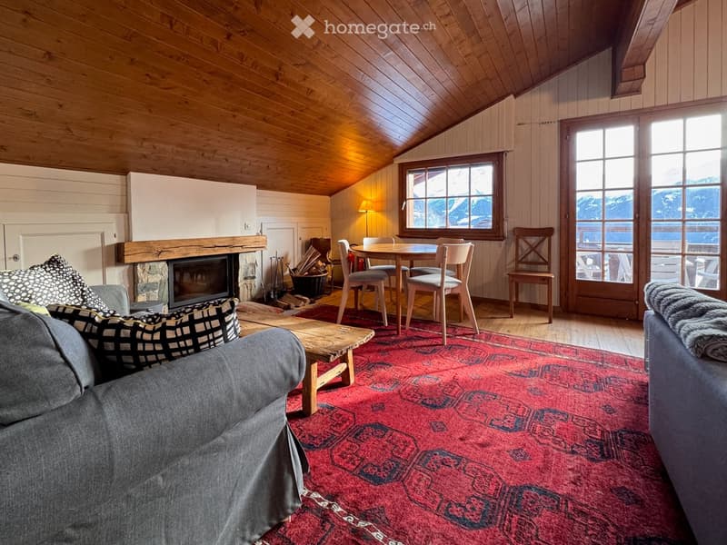 Cozy living room with wooden beams, fireplace, large windows, and a dining area.