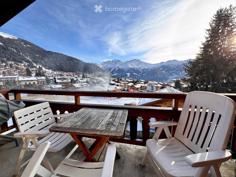 Balcony with wooden table and white chairs, overlooking a snowy mountain village.