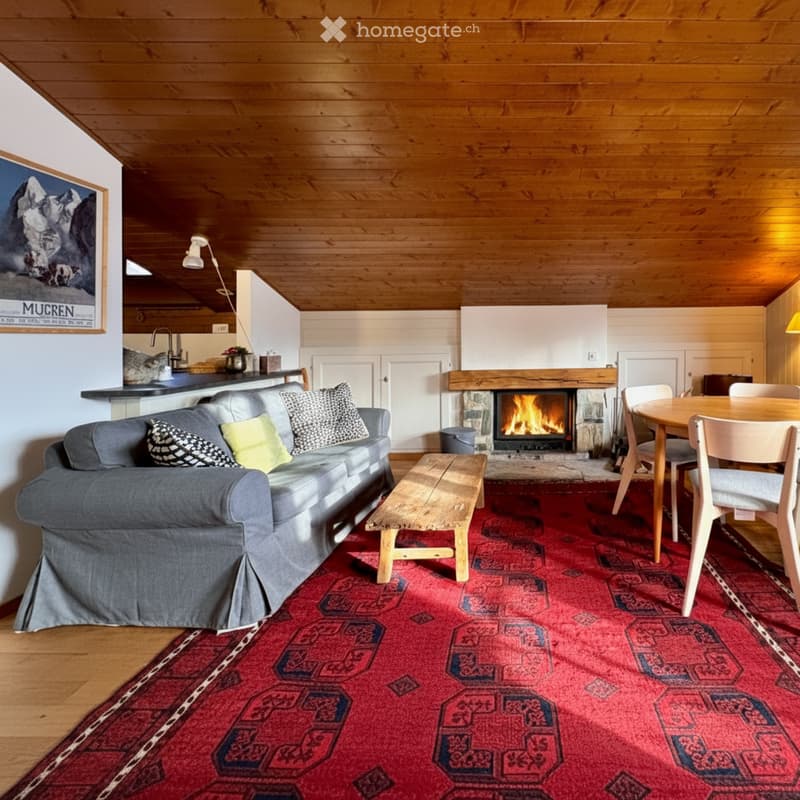 Cozy living room with wooden beams, a fireplace, a grey sofa, a wooden coffee table, and a dining area with a table and chairs.