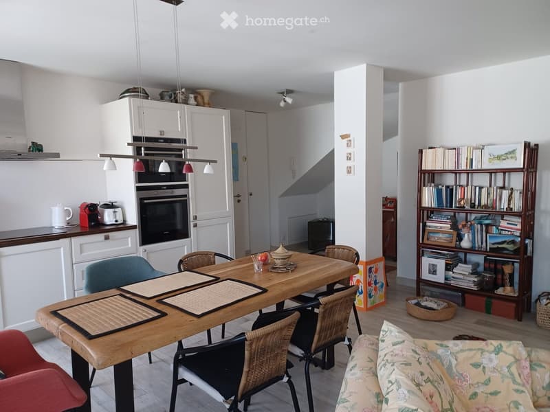 Open-plan living and dining area with a floral-patterned sofa, wooden dining table with placemats, wicker chairs, and a modern kitchen featuring white cabinets, an oven, and various appliances.