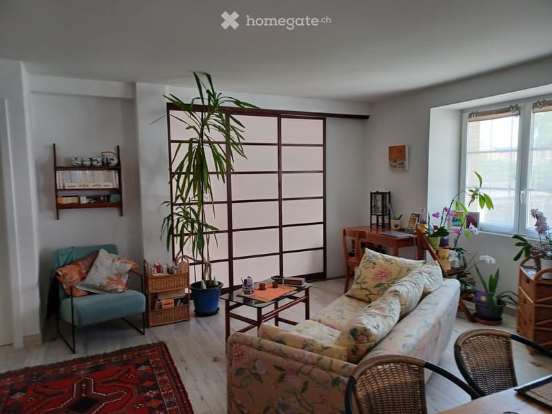A cozy living room with a floral-patterned sofa, a teal armchair, a coffee table, a large potted plant, a shelf with books and decorative items, and a small desk with a laptop and plants.