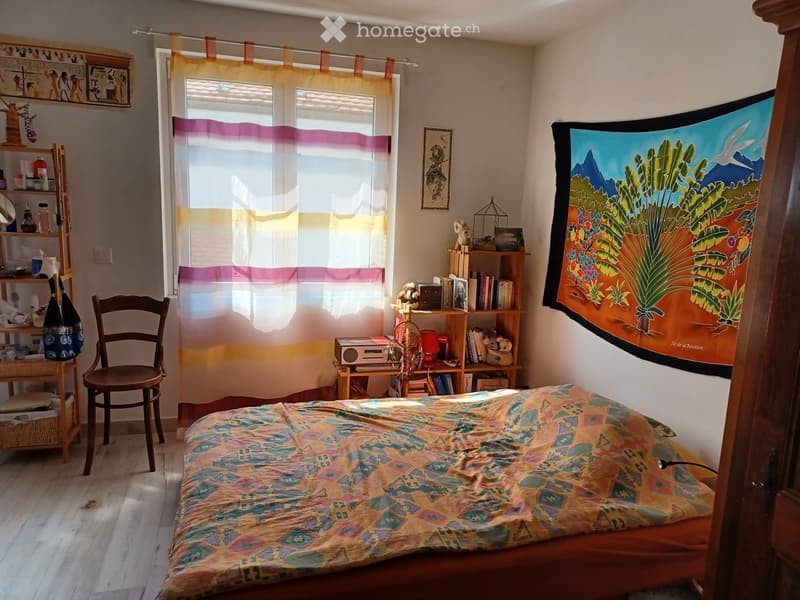 Cozy bedroom with wooden floor, large window with colorful curtains, bed with patterned bedspread, wooden chair, shelves with books and decorative items, and a colorful tapestry on the wall.