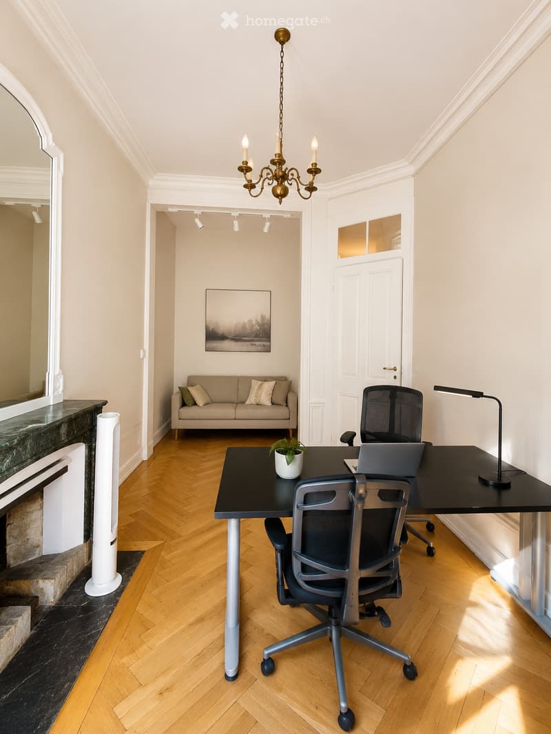 A modern, well-lit room with a wooden floor, a black desk with a laptop, a comfortable chair, a small sofa with pillows, a chandelier, and a potted plant. The room features a fireplace with a mirror above it and a view into another room with a painting on