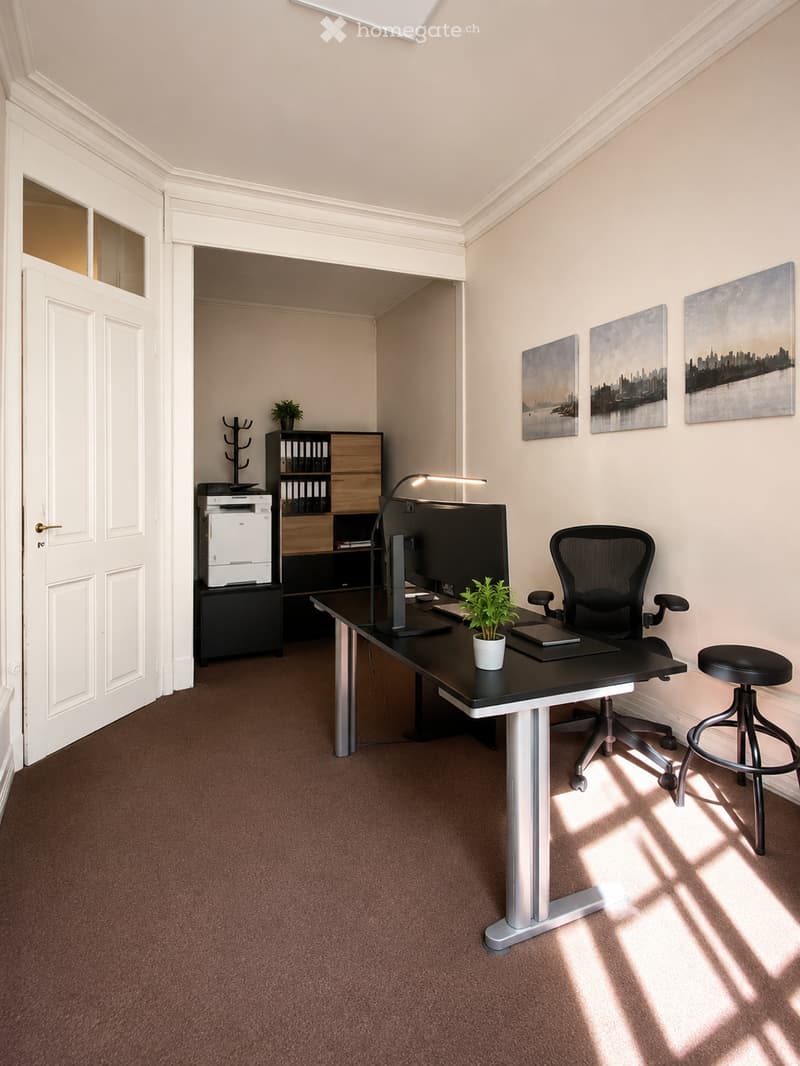 Modern office space with a black desk, ergonomic chair, stool, large screen, printer, cabinet, small plant, lamp, and artwork on the walls.