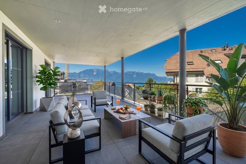 Modern balcony with a view of mountains, featuring a sofa, chairs, a table with drinks and snacks, and several potted plants.