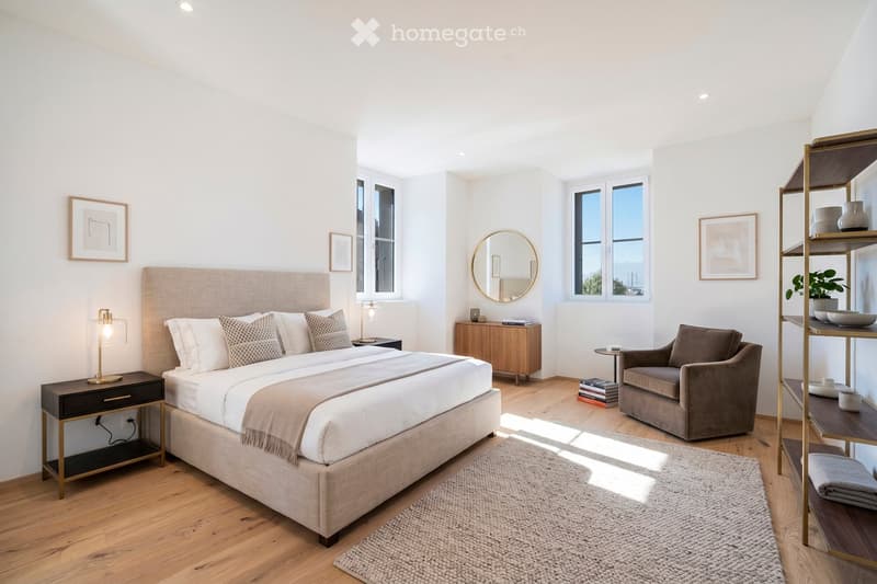 Modern bedroom with a large bed, nightstands, a cozy armchair, a wooden shelf, and a round mirror. The room features large windows, a woven rug, and decorative items on the shelves.