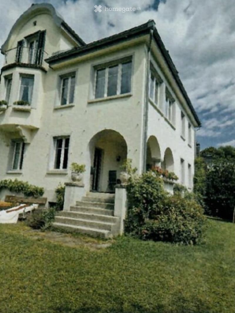 A two-story white house with arched doorways, multiple windows, and a staircase leading to the entrance, surrounded by greenery.