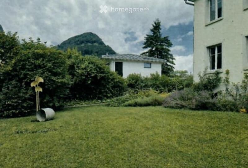 Well-maintained garden with green grass, bushes, and trees, featuring a scenic mountain view in the background.