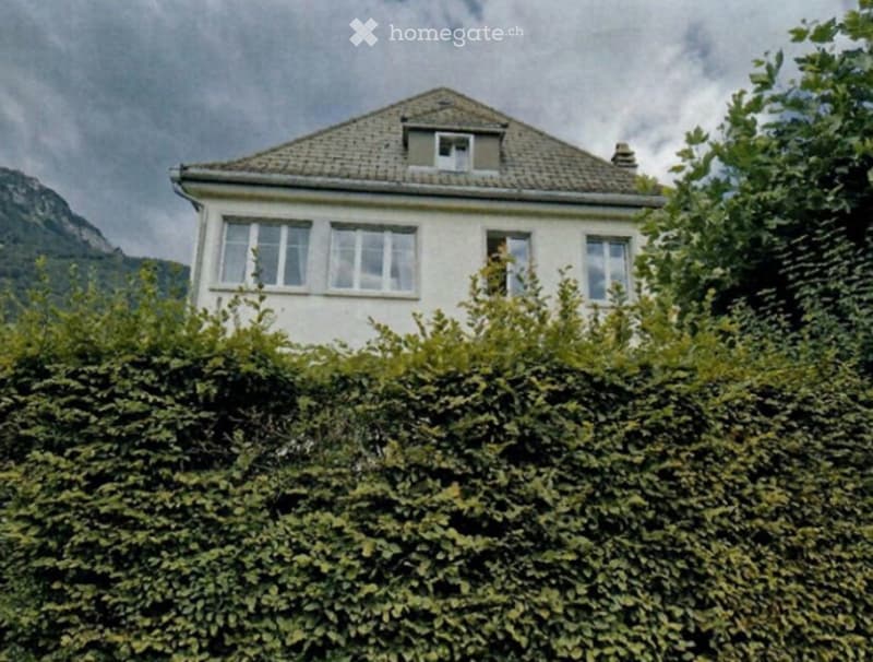 A two-story white house with multiple windows, surrounded by dense green foliage and a scenic mountain view in the background.