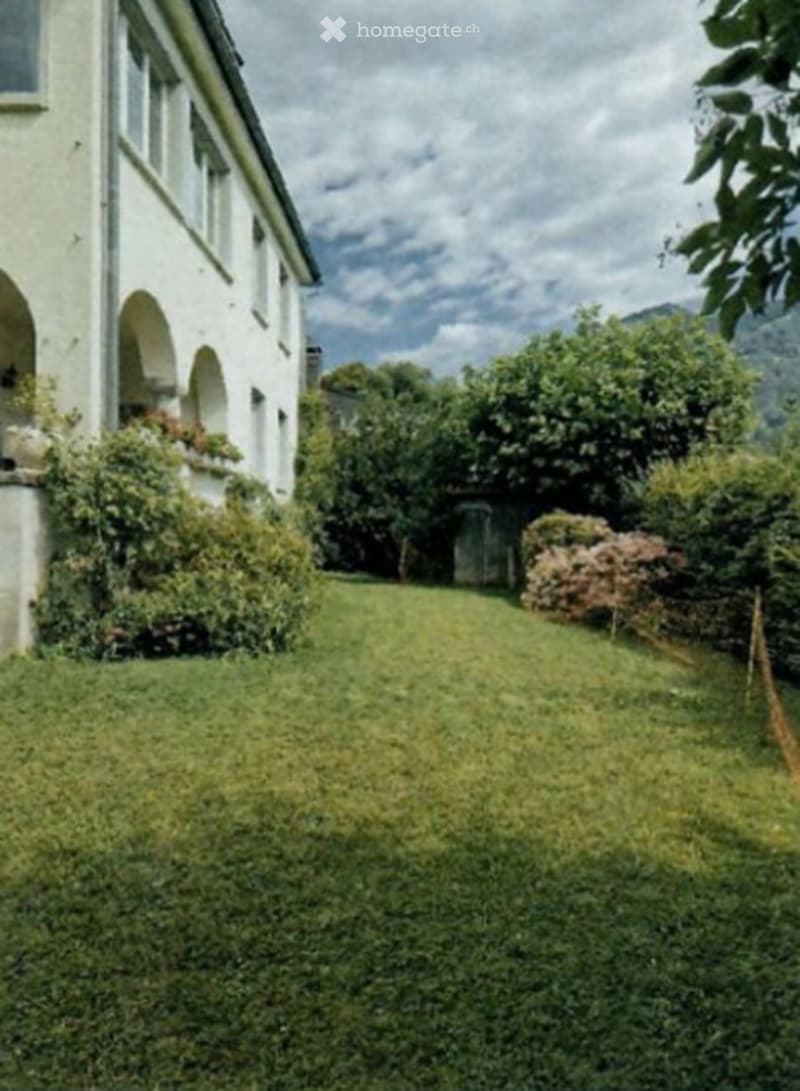 A well-maintained garden area with a lush green lawn, surrounded by a white building with arched windows and greenery. The background features trees and a scenic view of mountains under a partly cloudy sky.