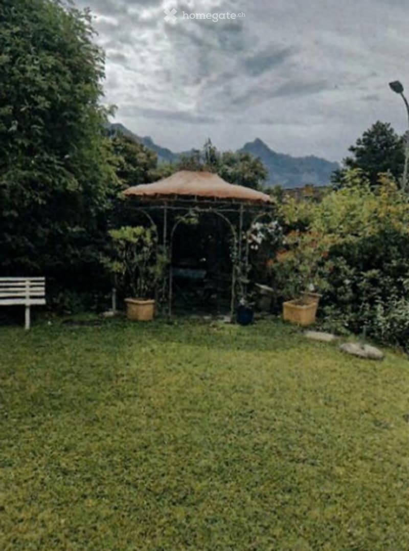A lush garden with a grassy lawn, a white bench, a gazebo with a thatched roof, and several potted plants. The background features trees and a mountainous landscape.