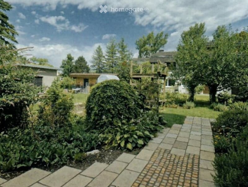 A well-maintained garden with a paved pathway, lush greenery, and a wooden deck area. The background shows a house and additional greenery.
