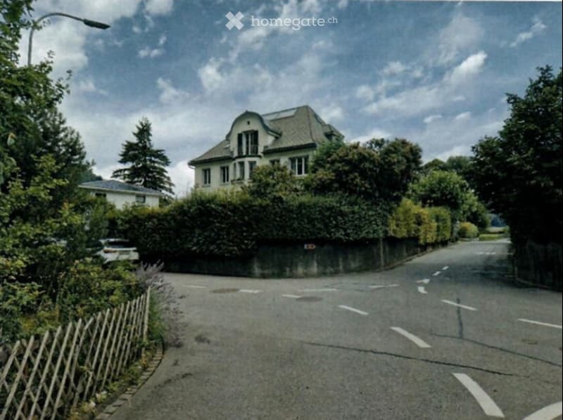 A large, multi-story house with a pitched roof and multiple windows, surrounded by greenery and a low hedge. The house is partially obscured by trees and a wooden fence along the street.