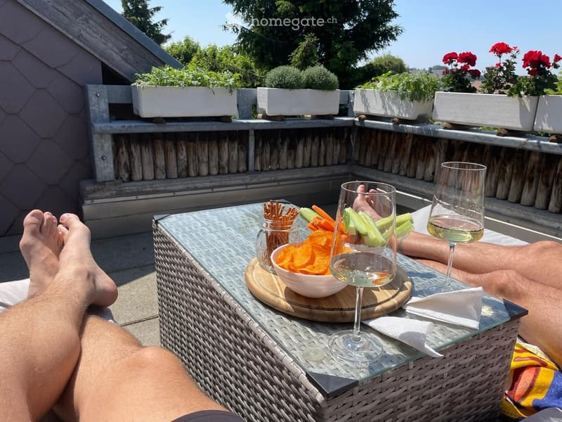 A sunny balcony with a glass-topped wicker table, snacks, and wine glasses, surrounded by potted plants and flowers.