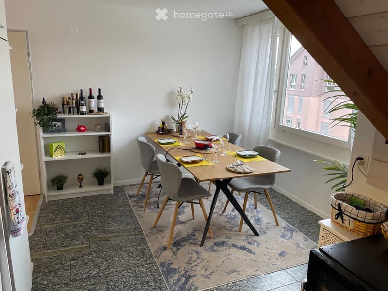 Modern dining area with a wooden table, six chairs, wine bottles, and decorative items on shelves.