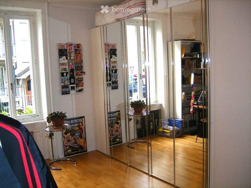 A modern, well-lit room with wooden flooring, large mirrors, a small round table with a potted plant, posters on the wall, and a desk area with shelves.