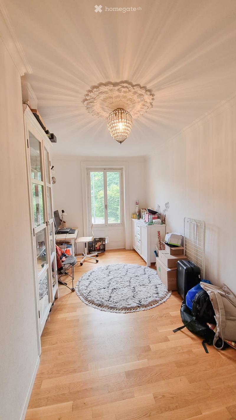 Bright room with wooden flooring, large window, chandelier, desk with laptop, chair, cabinet, and luggage near the wall.