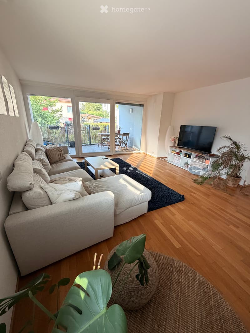 Bright living room with wooden flooring, large sectional sofa, coffee table, TV on a shelf, and plants. Large sliding glass doors lead to a balcony.