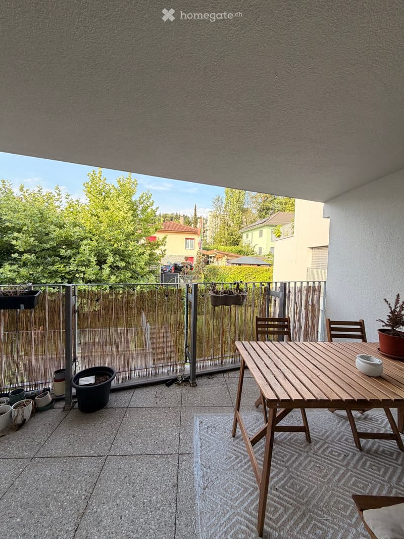 Balcony with wooden table, chairs, and potted plants, overlooking a green area with trees and houses.