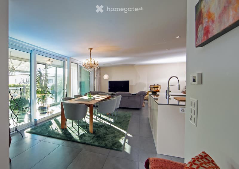 Open-plan living and dining area with large glass doors leading to a balcony, a sofa, dining table with chairs, television, chandelier, floor lamp, and a modern kitchen area with a faucet.