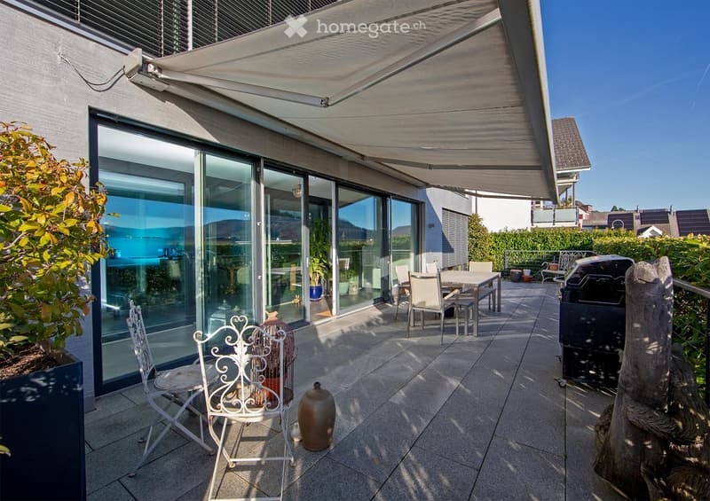 A modern, covered balcony with large glass sliding doors leading to an indoor space. The balcony features a dining table with chairs, a grill, and decorative elements like vases and a wooden sculpture.