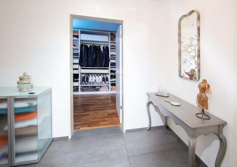 A well-lit hallway with a glass-fronted cabinet, a small table with a decorative item, and an open closet with neatly arranged clothing and shoes.