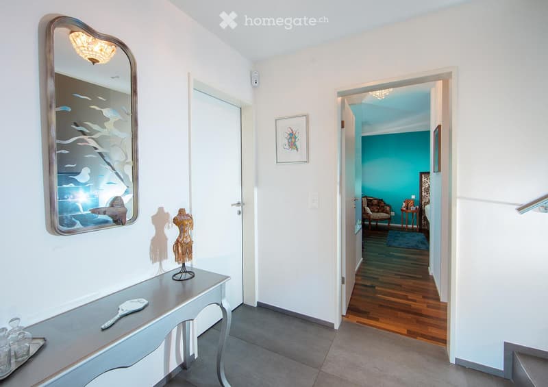A bright hallway with white walls, a decorative mirror, a small table with decorative items, and an open doorway leading to a room with teal walls and wooden flooring.