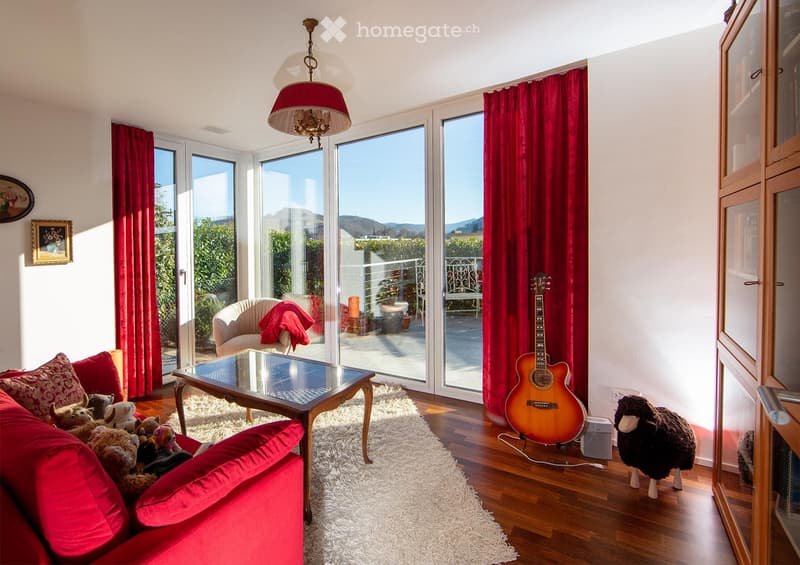 Bright living room with large windows and a view, red sofa with stuffed animals, glass coffee table, guitar, sheepskin, and a hanging lamp.