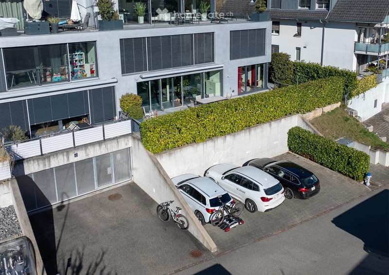 Modern multi-story residential building with large windows, balconies, and a parking area with several cars and bicycles.
