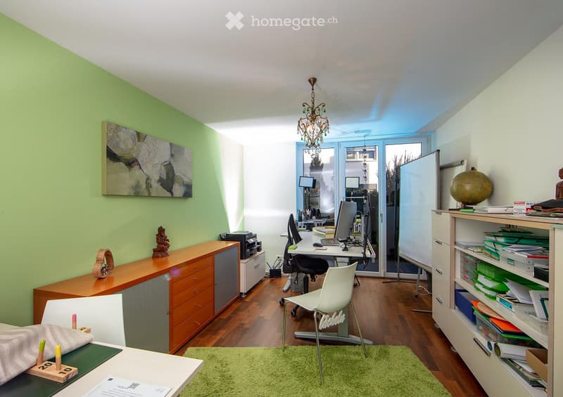 Office space with a wooden floor, green accent wall, white desk, laptop, chair, cabinet, shelf, globe, and artwork.
