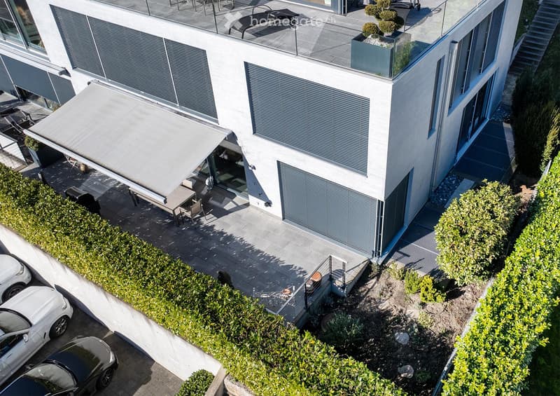 Modern two-story building with a rooftop terrace, outdoor seating, and a covered patio area. The building features large windows and a sleek design. There is a parking area with several cars visible.