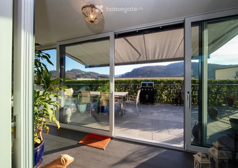 Spacious balcony with large glass sliding doors, outdoor dining set, grill, and scenic mountain view.