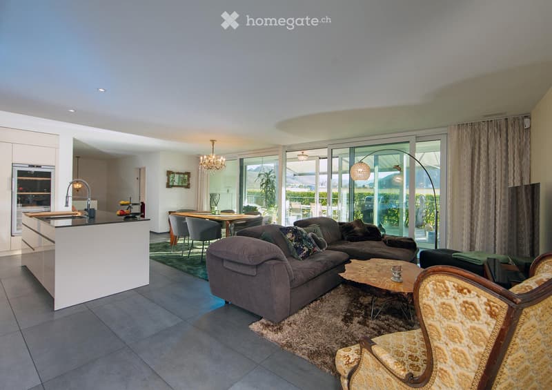 Open-plan living and dining area with a grey sectional sofa, coffee table, armchair, dining table with chairs, and a modern kitchen island with a sink and faucet. Large windows offer a view of the garden.